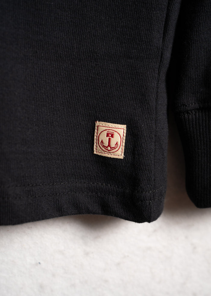 Close-up of a black sweater's hem with a small beige patch featuring a red anchor design, set against a light background.