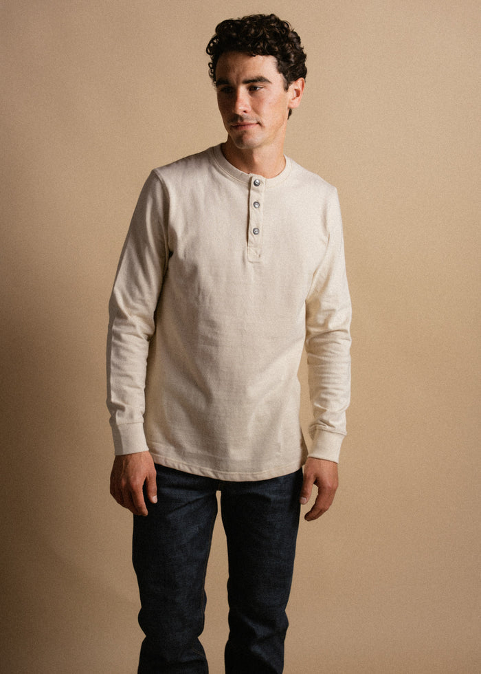 Man in a cream henley shirt and dark jeans stands against a beige background, looking to the side with a neutral expression.