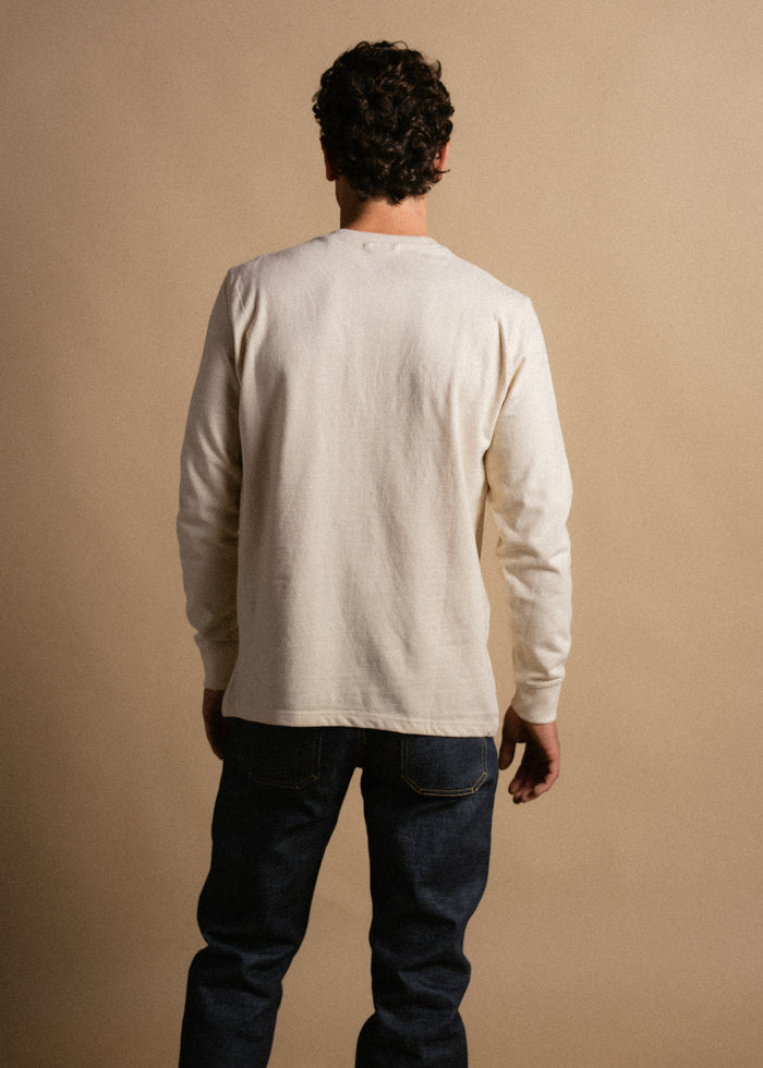 Man in a white long-sleeve shirt and jeans stands facing away against a beige background, highlighting casual fashion style.
