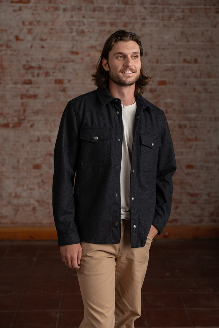 Man in a dark jacket and beige pants stands smiling against a rustic brick wall, exuding a casual, relaxed style.
