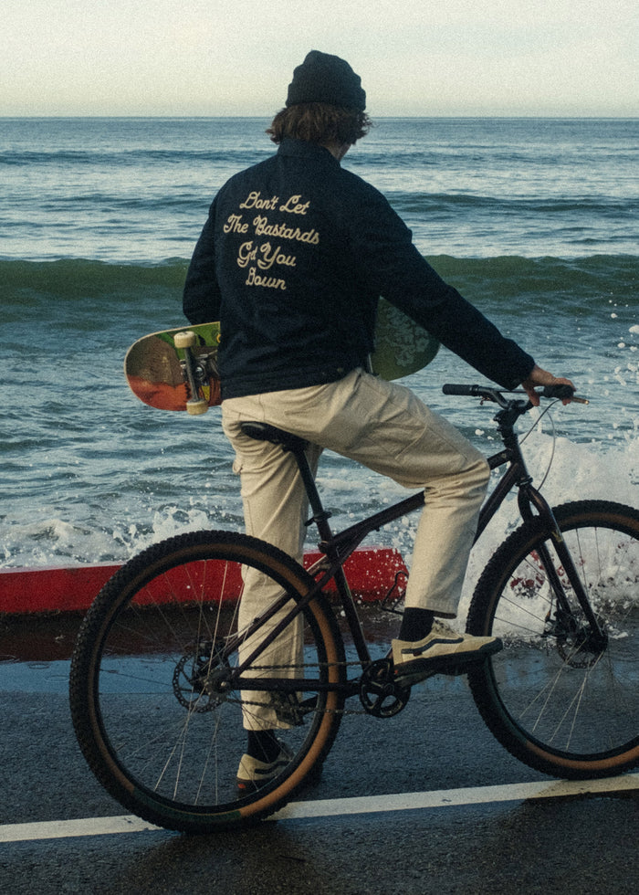 Cyclist by ocean, holding skateboard. Jacket reads "Don't Let The Bastards Get You Down." Waves crash on the shore.