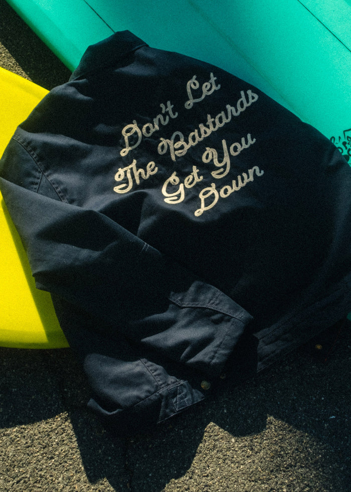 A navy jacket with "Don't Let The Bastards Get You Down" text lies on yellow and turquoise surfboards under sunlight.
