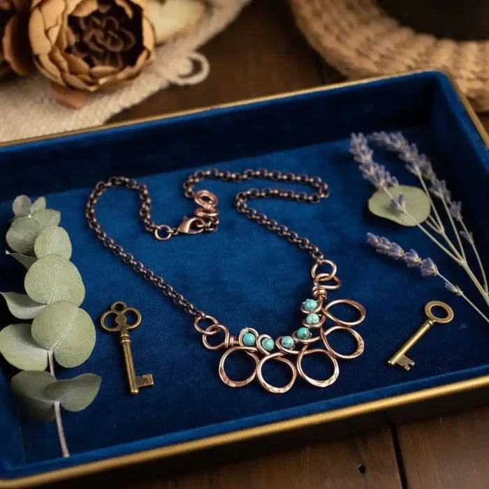 Seen on Ashley Liao Copper Turquoise Wire Wrapped Necklace displayed on a blue velvet tray with decorative elements.