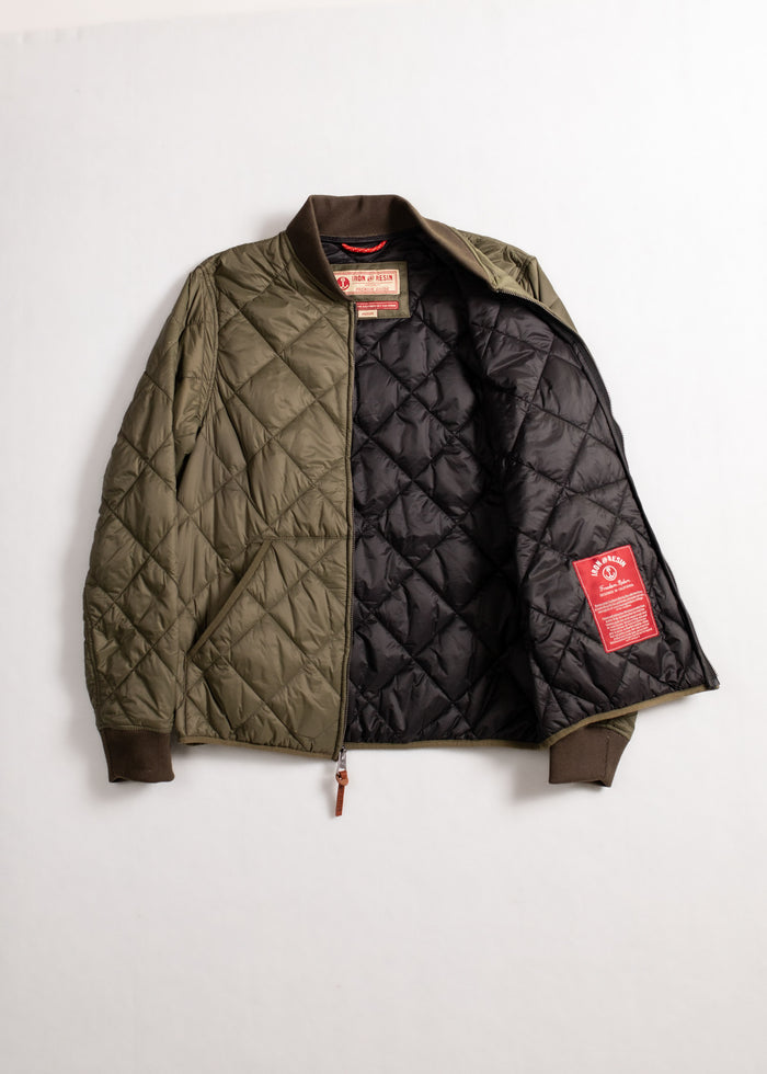 Olive quilted jacket with brown cuffs, open to show black lining and red label. Simple, stylish outerwear on a plain background.