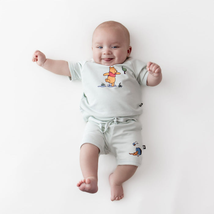 Smiling infant laying on a white surface wearing the Short Sleeve Jogger Set in Winnie the Pooh on Dew