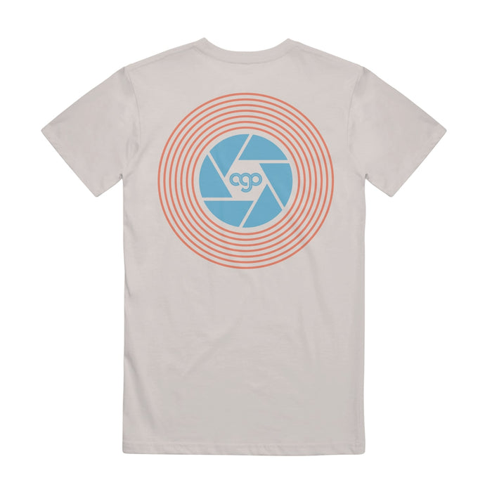 Back of sand colored short sleeve shirt with a camera shutter design on the back drawn in orange and blue. Inside the shutter there is small blue text that says AGO.