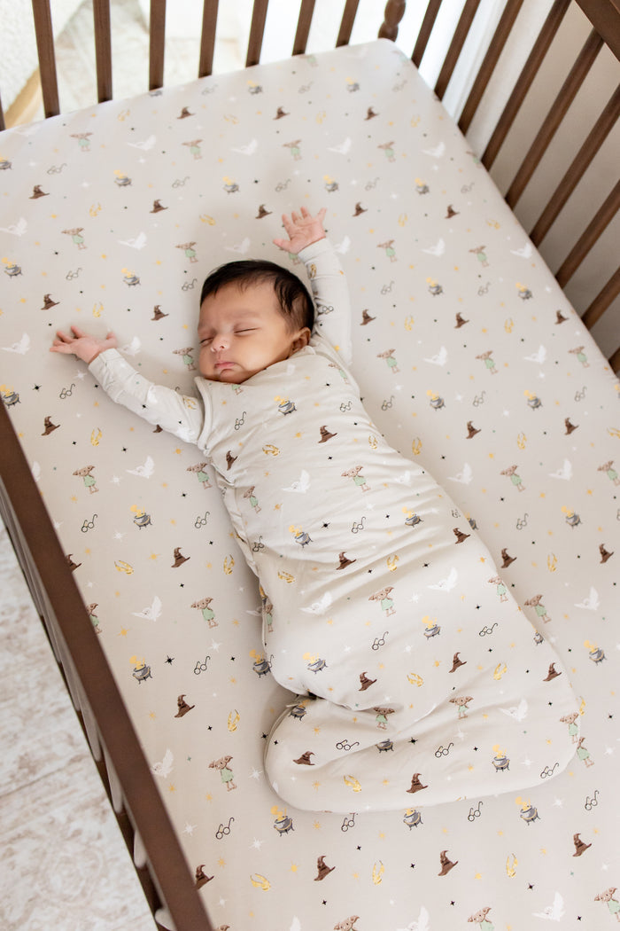 Baby wearing beige sleep bag with Harry Potter prints on it asleep in a crib on a similar print crib sheet