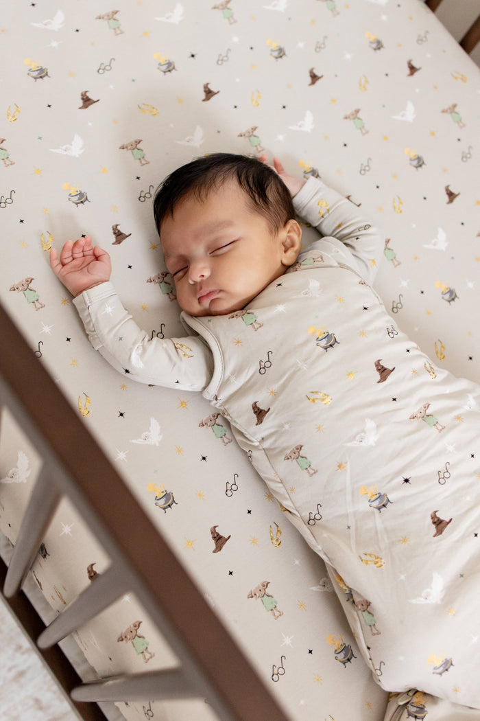 Baby wearing a beige Harry Potter patterned sleep bag sleeping on a similar print crib sheet.
