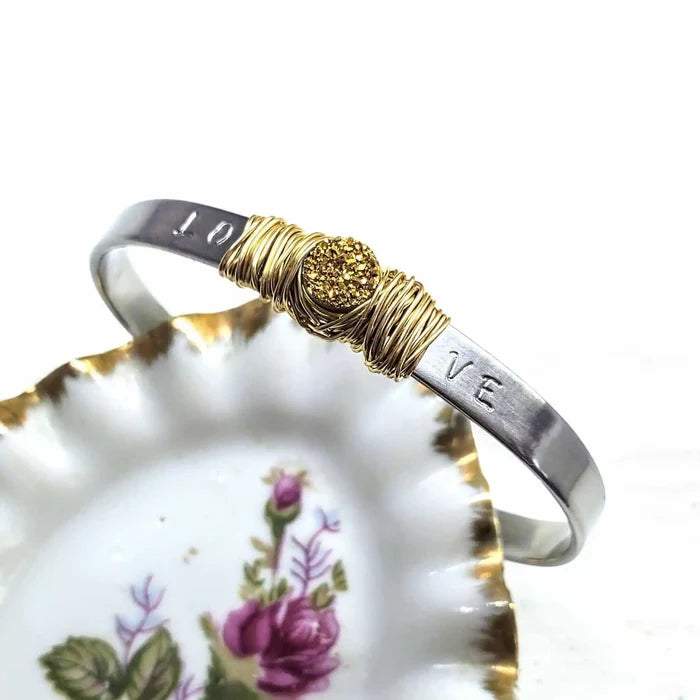 Stainless Steel LOVE Stamped Wire Wrapped Gold Druzy Cuff displayed on a decorative plate with floral pattern.
