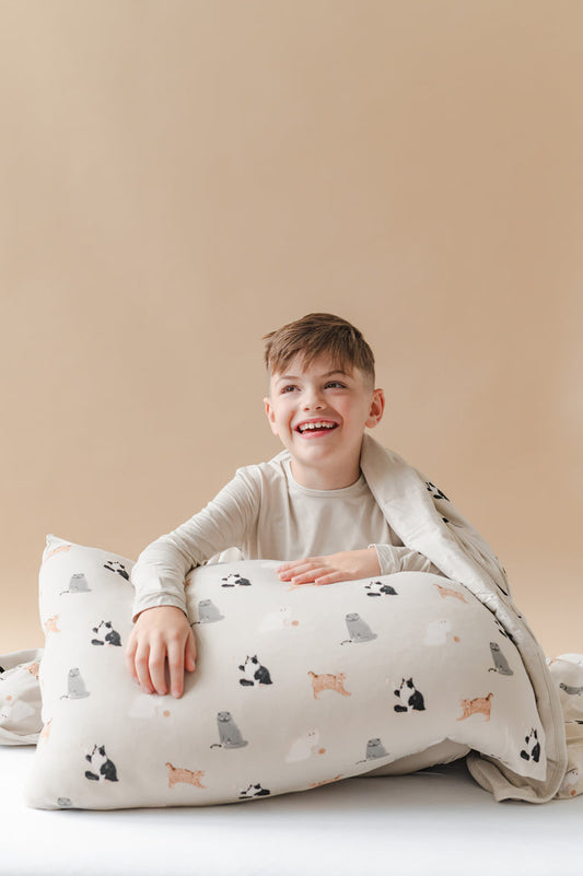 Child sitting on a bed with a cat patterned pillow and blanket against a beige wall.