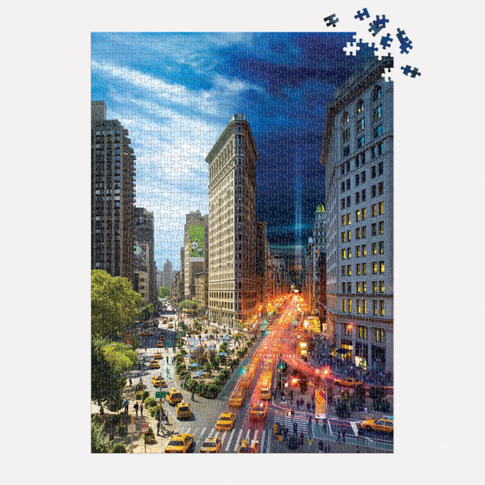 The Stephen Wilkes Day To Night Flatiron 1000 Piece Puzzle features a cityscape with the Flatiron Building, yellow taxis, and busy streets transitioning from day on the left to night on the right, with some pieces missing in the top right corner.