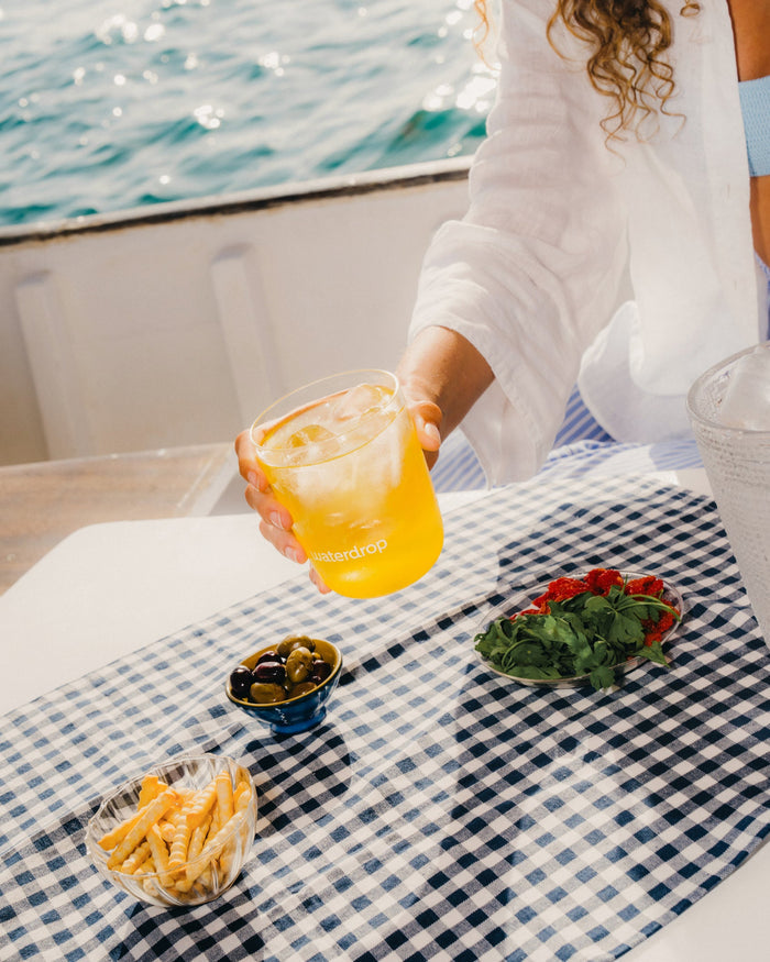 Person holding a glass of ORANGE, a sugar-free citrus drink, over a table with food, including french fries and olives.