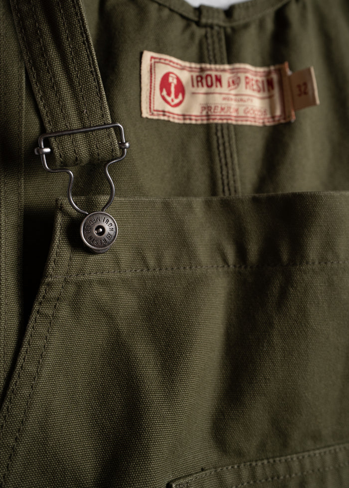 Close-up of green overalls with a metal button and "Iron and Resin" label, showcasing rugged design and premium craftsmanship.