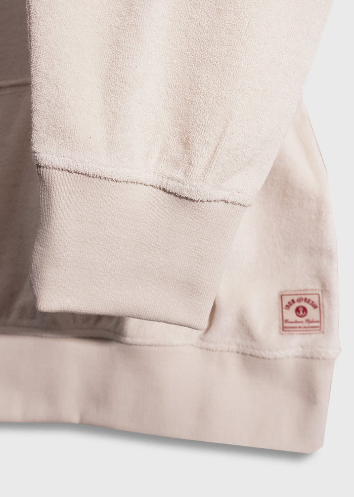 Close-up of a beige sweatshirt sleeve with ribbed cuff and a small red label on the hem, displaying texture and stitching details.