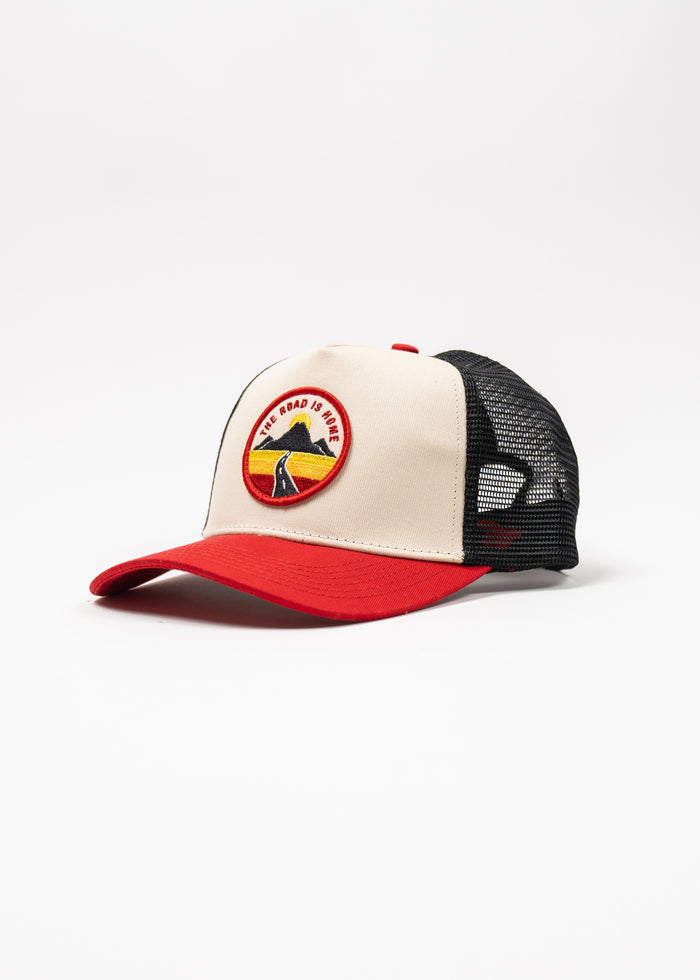 Trucker cap with red brim, beige front, black mesh, and patch reading "The Road is Home" featuring a mountain and sunset.
