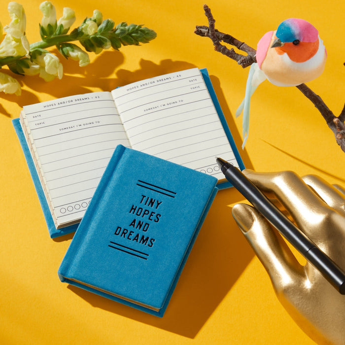 A small blue Brass Monkey Tiny Hopes and Dreams Tiny Diary with gilded-edge pages rests on a yellow surface beside a matching open notebook, a gold hand holding a black pen, an artificial bird on a branch, and greenery.