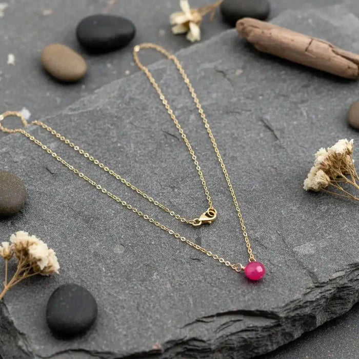 Tiny Hot Pink Chalcedony Gold Filled Necklace displayed on a slate stone with pebbles and dried flowers.