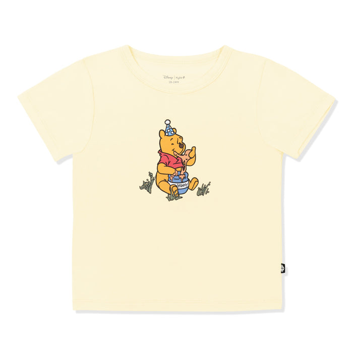 Light Yellow basic tee with the graphic of winnie the pooh sitting in grass eating honey out of a honey pot in the center of the shirt