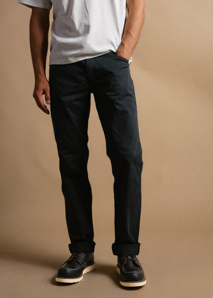 Man in a white t-shirt and black pants stands against a beige background, wearing dark leather shoes with white soles.