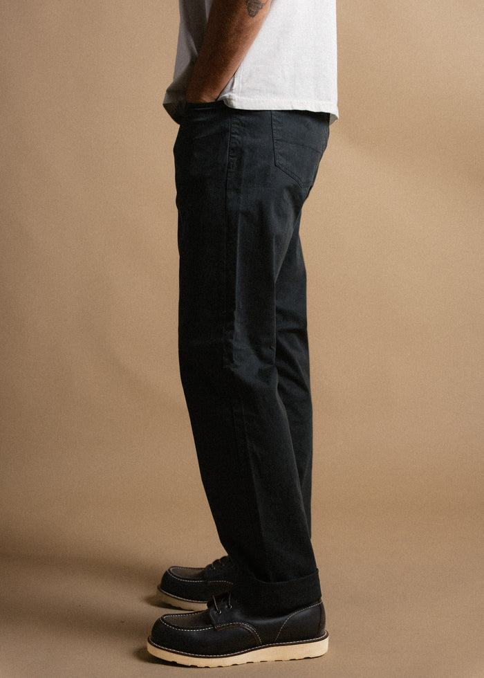 Man in black pants and brown boots stands against a beige background, showcasing casual style and footwear.