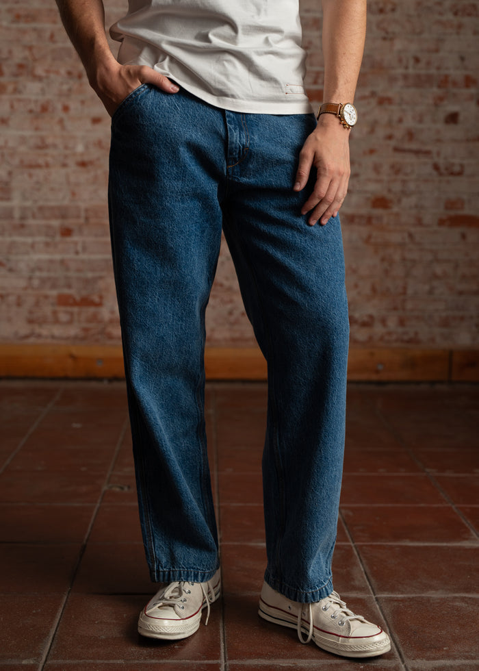 Person in casual jeans and white sneakers stands on a tiled floor, wearing a white shirt and watch, with a brick wall backdrop.