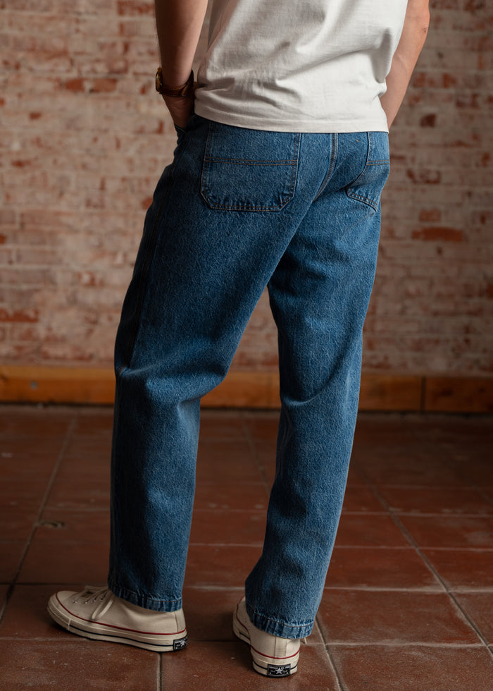 Person in blue jeans and white sneakers stands on a tiled floor, facing a brick wall, hands in pockets, wearing a white shirt.