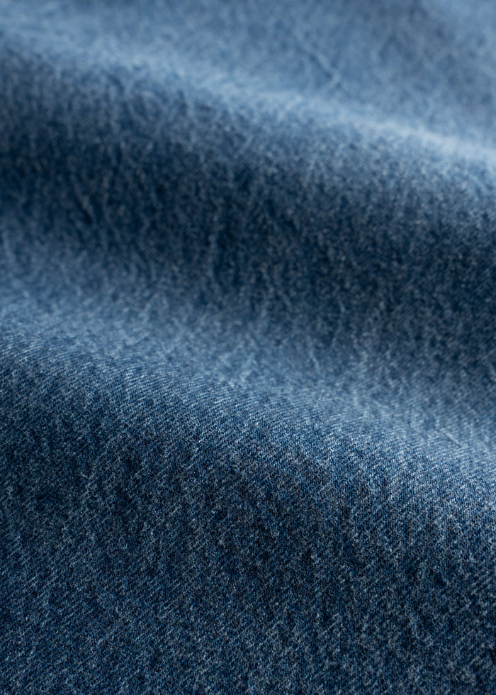Close-up of blue denim fabric with a textured surface, showing subtle variations in color and weave, creating a wavy pattern.