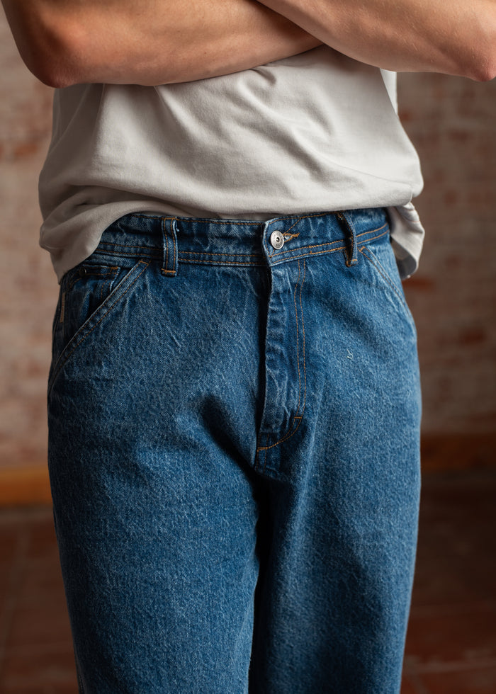 Person in blue jeans and a white shirt stands with arms crossed, showing casual style against a blurred brick background.