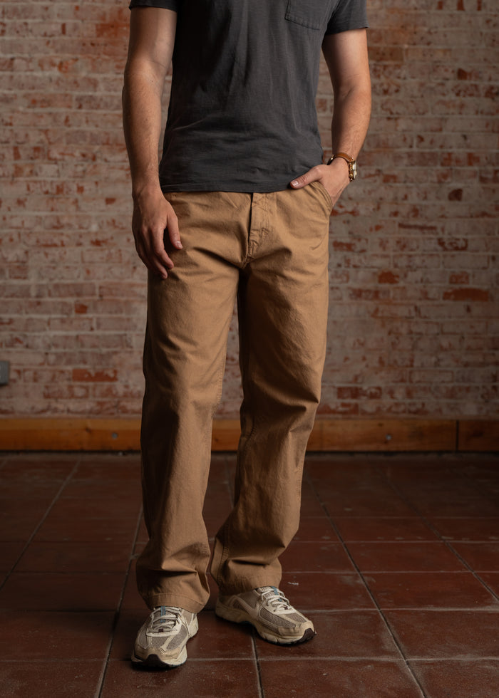 Man in a black t-shirt and khaki pants stands against a brick wall, hands in pockets, wearing beige sneakers on a tiled floor.