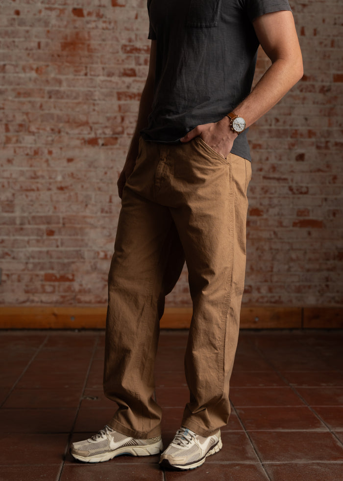 Man in casual outfit with brown pants and gray shirt, wearing a watch, stands against a brick wall, hands in pockets.