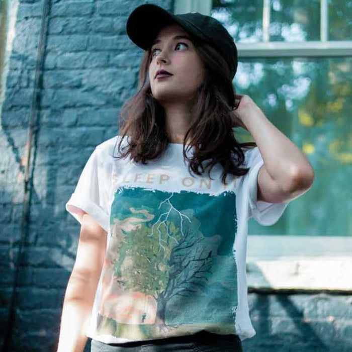 White short sleeve shirt with orange text that says SLEEP ON IT. Below the text, there is a drawing of a tree, on one side there is someone on a swing and the tree is alive, but on the other the tree is dead and has been struck by lightning. Below the drawing there is smaller orange text that says PRIDE & DISASTER. An individual is modeling the shirt and standing in front of a blue brick building.