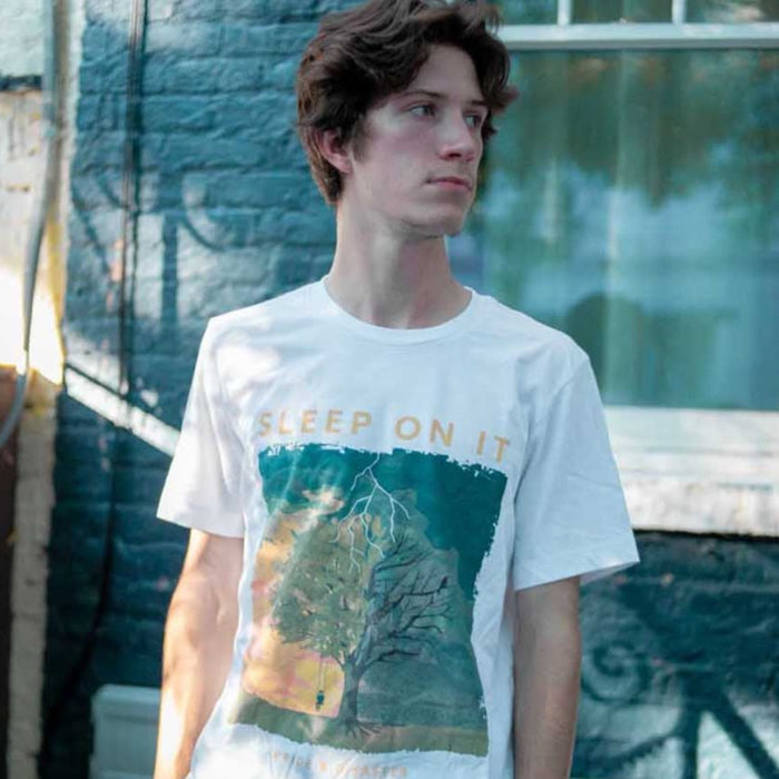 White short sleeve shirt with orange text that says SLEEP ON IT. Below the text, there is a drawing of a tree, on one side there is someone on a swing and the tree is alive, but on the other the tree is dead and has been struck by lightning. Below the drawing there is smaller orange text that says PRIDE & DISASTER. An individual is modeling the shirt and standing in front of a blue brick building.