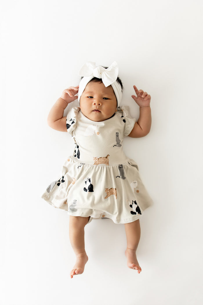 Infant wearing the Twirl Bodysuit Dress in Fluffy Cat paired with a white bow laying on a white surface