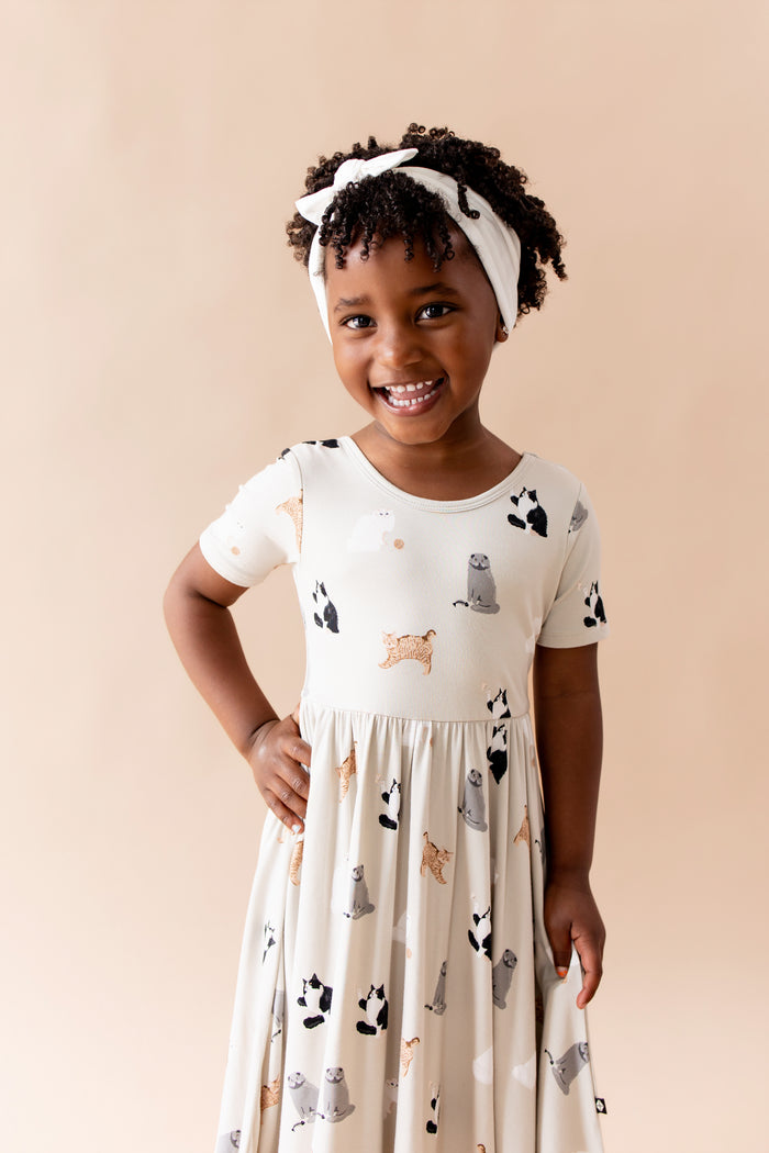 Smiling girl posing wearing the Twirl Dress in Fluffy Cat holding the skirt of the dress