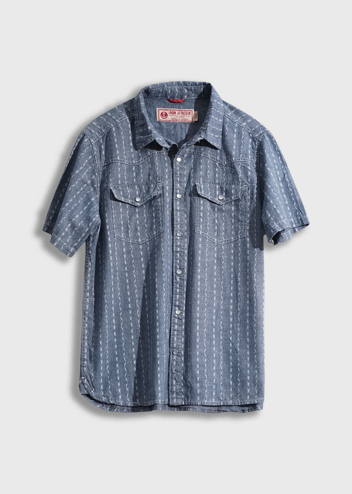 Blue short-sleeve denim shirt with white patterns, button front, and two chest pockets, hanging against a plain background.
