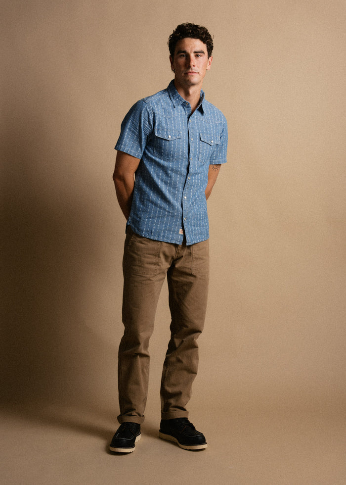 Man in a blue short-sleeve shirt and khaki pants stands against a beige background, hands in pockets, looking at the camera.