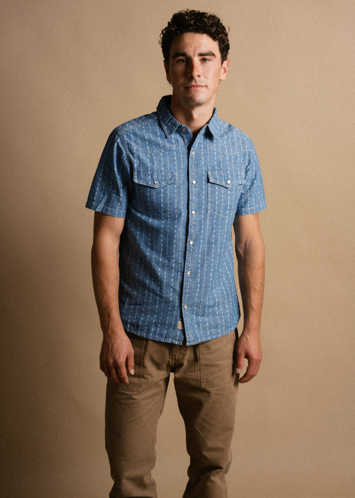 Man in a blue short-sleeve button-up shirt and brown pants stands against a beige background, looking directly at the camera.