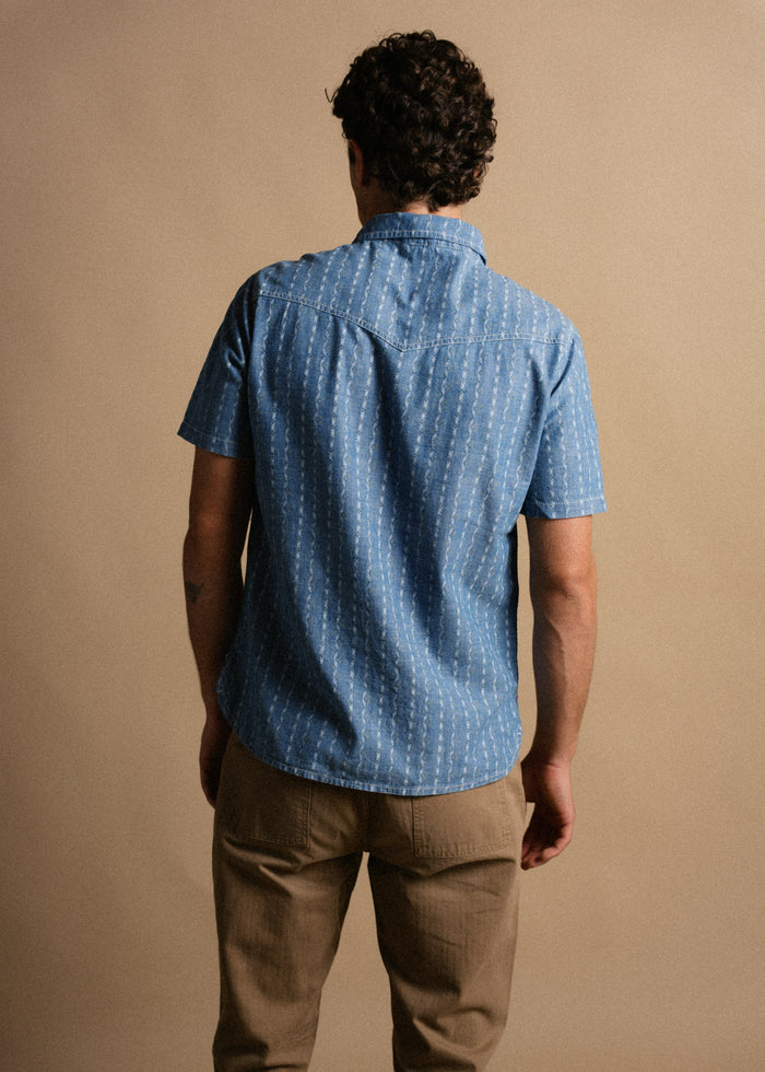 Man in blue patterned shirt and beige pants stands with back to camera against a neutral background, showcasing casual style.