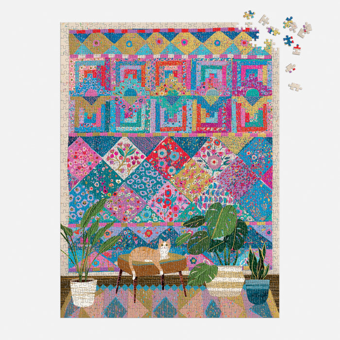 The Victoria Ball Quilters Corner 1000 Piece Foil Puzzle by Victoria Ball depicts a cat resting on a table amid potted plants, with an ornate quilt tapestry on the wall and some unfinished pieces in the top right corner.