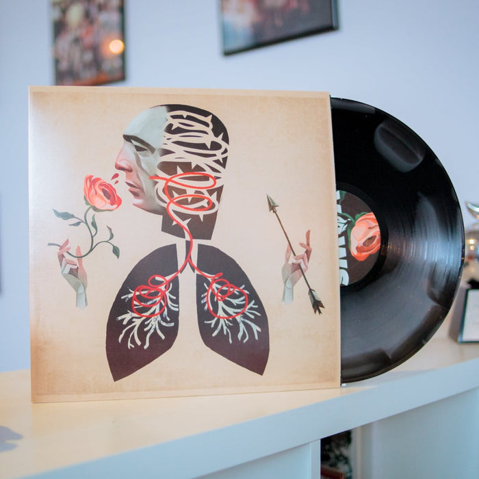 Silver and black marble swirl vinyl. The jacket is a faded brown color. The vinyl and jacket rest against a white table top in an office setting. The design is of a head with a distinct face. The head is connected to two lungs. There are hands on either side of the design. The right hand is holding a rose. The mask is smelling the rose, and red swirly lines are seen going from the nose to the lungs. The left hand holds an arrow.