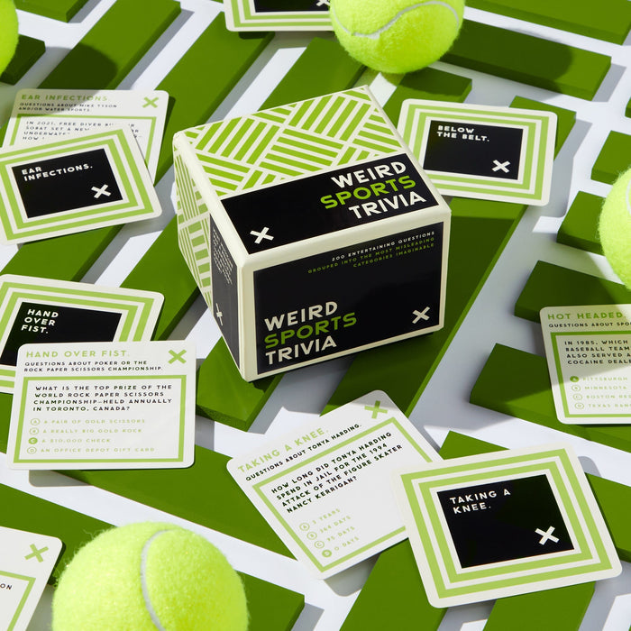 Brass Monkeys Weird Sports Trivia box sits on a green and white surface, surrounded by several cards and yellow tennis balls. The cards feature obscure sports facts and unusual sports-related questions.