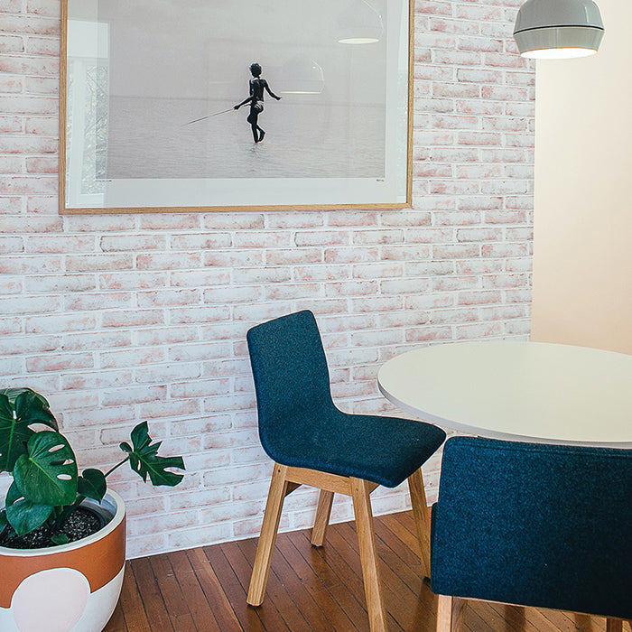 Tempaper's Whitewashed Brick Peel And Stick Wallpaper shown behind a plant and white table with blue chairs. #color_whitewashed