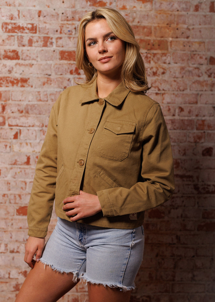 Woman in tan jacket and denim shorts stands against a brick wall, looking to the side with a thoughtful expression.