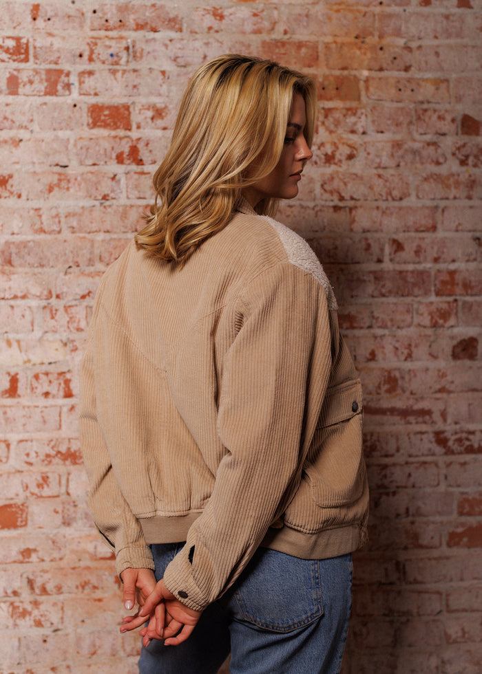 Blonde woman in a tan corduroy jacket and jeans stands against a rustic brick wall, hands clasped behind her back, looking sideways.