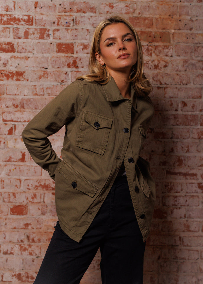 Blonde woman in an olive jacket poses confidently against a rustic brick wall, hands in pockets, exuding casual elegance.