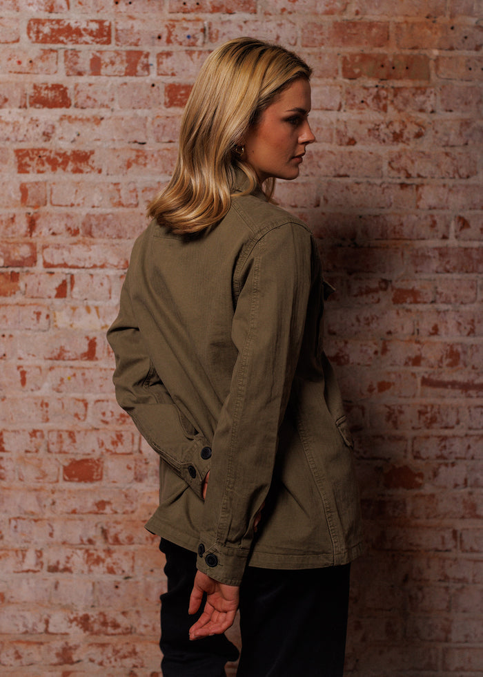 Woman in olive jacket stands against a textured brick wall, looking to the side with hands behind her back, blonde hair flowing.
