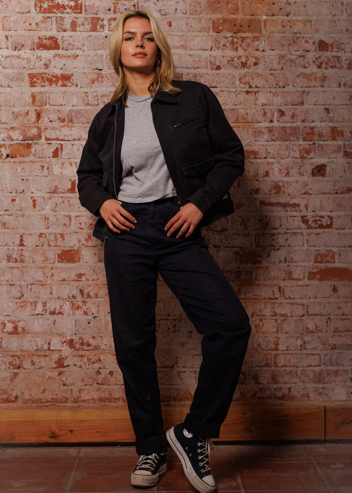 A person in a black jacket and jeans stands confidently against a rustic brick wall, wearing sneakers and a gray shirt.