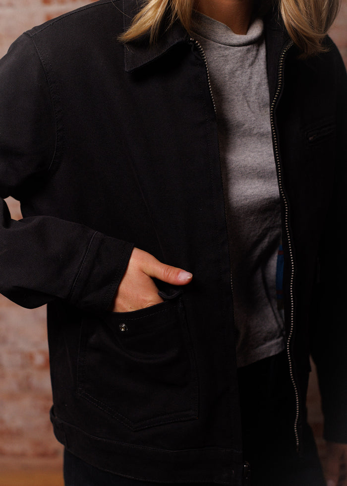 Person in a black jacket with hand in pocket, wearing a gray shirt. Brick wall background adds texture to the scene.