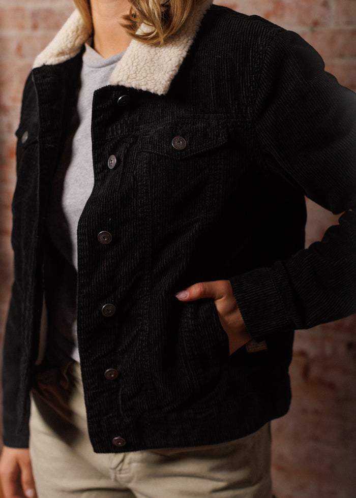 Person wearing a black corduroy jacket with a white fleece collar, hand in pocket, standing against a brick wall background.