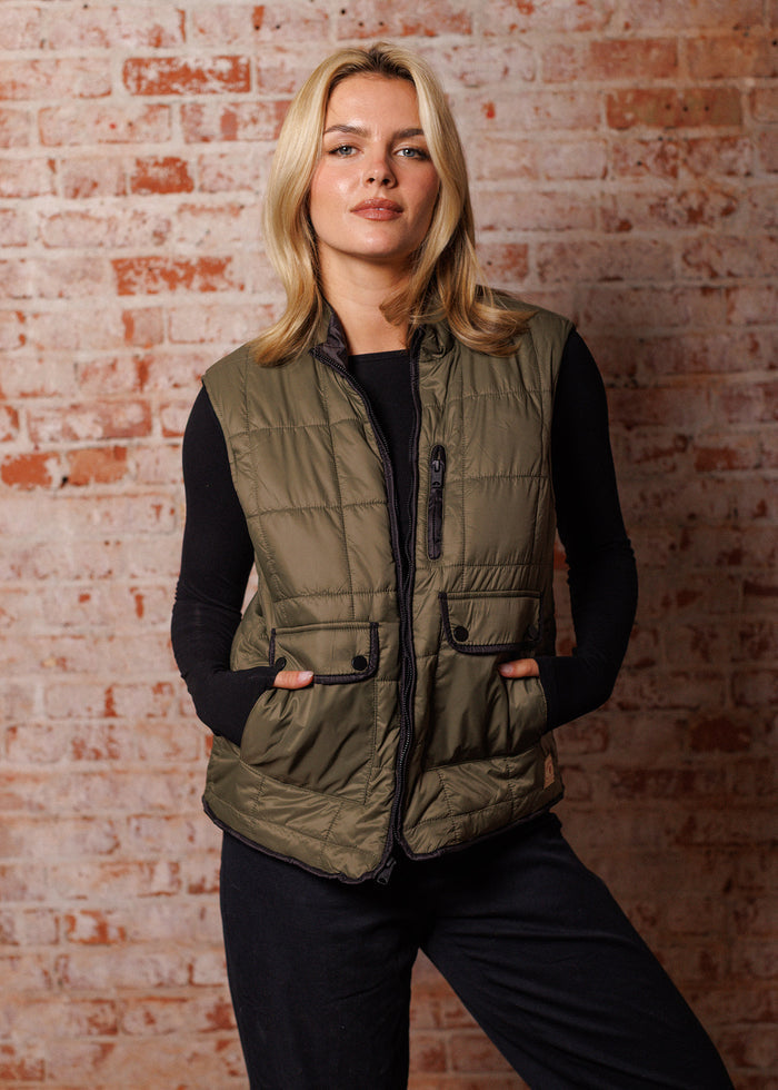 Blonde woman in olive quilted vest and black top stands confidently against a textured brick wall, hands in pockets.